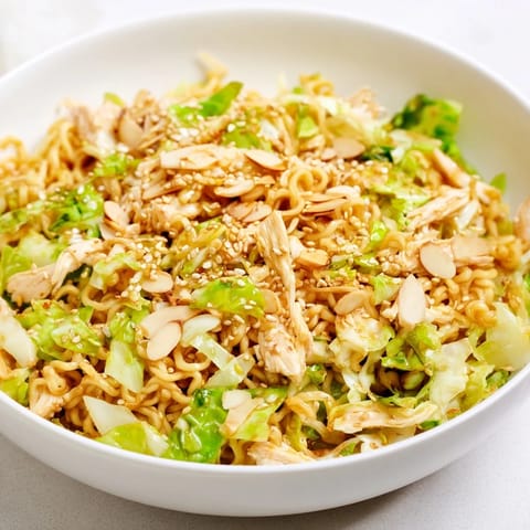 A bowl of crunchy ramen chicken salad garnished with toasted almonds, sesame seeds, and fresh cilantro beside a simple serving fork.